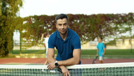 Portrait-Of-A-Handsome-Man-Coming-Closer-To-Camera-With-Racket-In-Hand,-Leaning-On-Net-And-Smiling-At-The-Camera-While-Spending-Time-With-His-Family-On-A-Tennis-Court