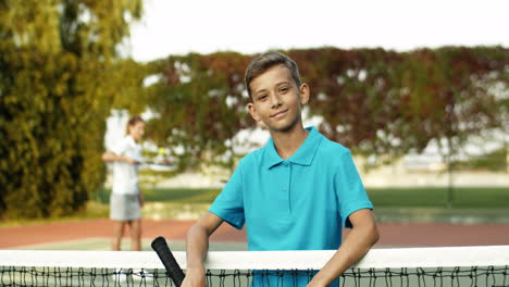Portrait-Of-A-Cute-Teen-Boy-With-Tennis-Racket-In-Hand-Leaning-On-Net-And-Smiling-Cheerfully-At-The-Camera