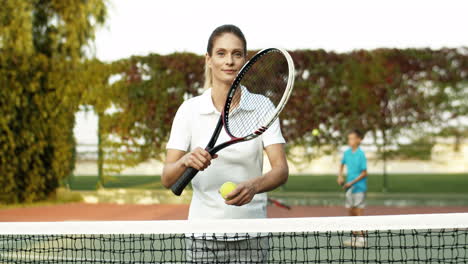 Portrait-Of-A-Beautiful-Woman-Coming-Closer-To-Camera-With-Racket-In-Hand,-Leaning-On-Net-And-Smiling-At-The-Camera-While-Spending-Time-With-Her-Family-On-A-Tennis-Court