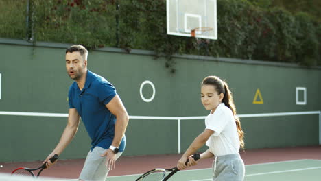 Man-Teaching-His-Little-Daughter-How-To-Play-Tennis-On-A-Summer-Day