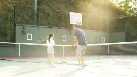 Man-Teaching-His-Little-Daughter-How-To-Play-Tennis-On-A-Summer-Day-1