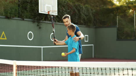 Man-Teaching-His-Teen-Son-How-To-Hit-Ball-With-Racket-On-A-Tennis-Court-On-A-Summer-Day