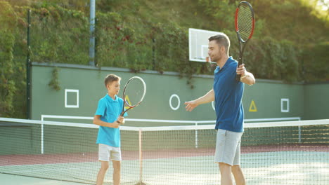 Man-Teaching-His-Teen-Son-How-To-Play-Tennis-On-An-Outdoor-Court-In-Summer-And-Then-Giving-High-Five
