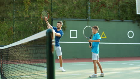Man-Teaching-His-Teen-Son-How-To-Play-Tennis-And-Then-Giving-High-Five-On-An-Outdoor-Court-In-Summer