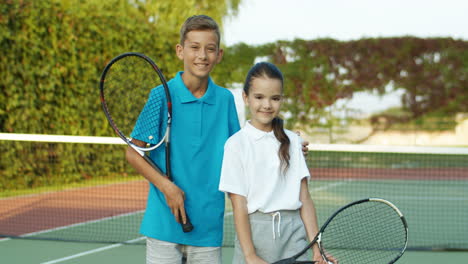 Portrait-Of-Cute-Brother-And-Sister-Holding-Rackets-And-Smiling-Cheerfully-At-The-Camera-While-Standing-On-An-Outdoor-Tennis-Court