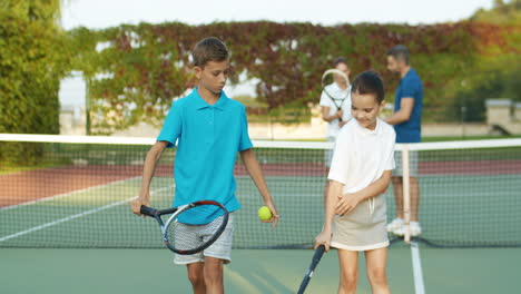 Older-Brother-Teaching-Younger-Sister-How-To-Play-Tennis-At-Sport-Court-On-Summer-Day-While-Their-Parents-Talking-In-The-Background