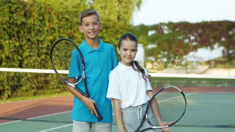 Portrait-Of-Cute-Brother-And-Sister-Holding-Rackets-And-Smiling-Cheerfully-At-The-Camera-While-Standing-On-An-Outdoor-Tennis-Court-1