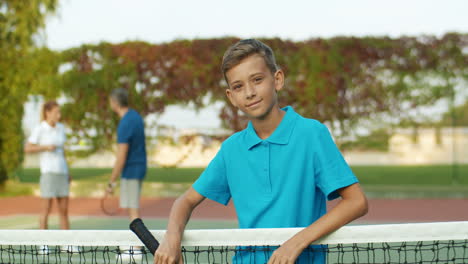 Porträt-Eines-Süßen-Teenagers-Mit-Tennisschläger-In-Der-Hand,-Der-Sich-Am-Netz-Lehnt-Und-Fröhlich-In-Die-Kamera-Lächelt-1