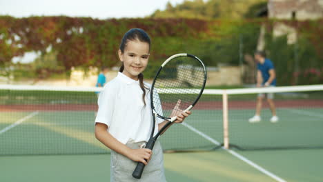 Portrait-Of-Cute-Little-Girl-Holding-Racket-And-Smiling-Cheerfully-At-The-Camera-While-Standing-On-An-Outdoor-Tennis-Court-1