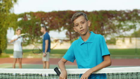 Portrait-Of-A-Tired-Teen-Boy-With-Racket-Leaning-On-Net-And-Smiling-At-The-Camera-On-A-Tennis-Court