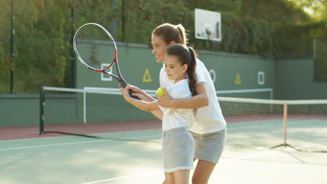 Loving-Woman-Teaching-Her-Cute-Little-Daughter-How-To-Play-Tennis-At-Sport-Court-On-A-Summer-Day