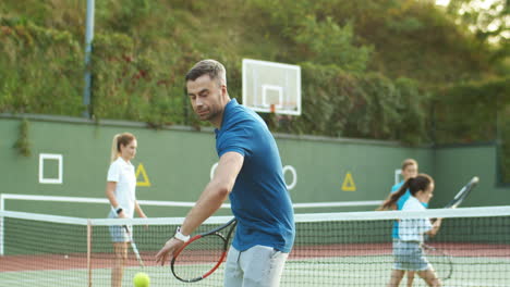 Schöner-Mann-Trainiert-Und-Schlägt-Ball-Mit-Schläger,-Während-Seine-Familie-Im-Hintergrund-Tennis-Spielt