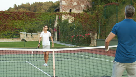 Rear-View-Of-Sporty-Man-Playing-Tennis-With-Blonde-Woman-At-Outdoor-Court-On-A-Summer-Day