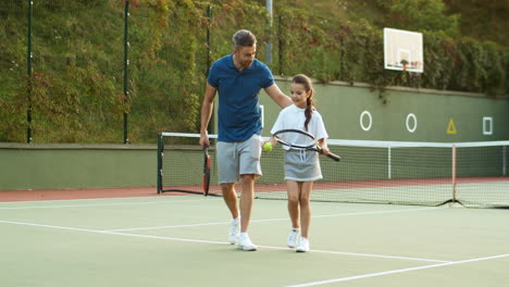 Liebevoller-Vater-Und-Glückliche-Kleine-Tochter,-Die-Scherzen-Und-Gemeinsam-Den-Tennisplatz-Verlassen
