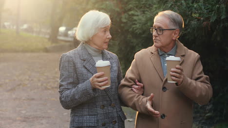 Elderly-Married-Couple-Walking-In-The-Park,-Talking-And-Drinking-Hot-Coffee-At-Sunset-In-Autumn