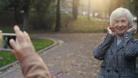 Elderly-Woman-With-Grey-Hair-And-Wearing-Coat-Posing-In-The-Park-While-Her-Husband-Taking-A-Photo-Of-Her-With-Smartphone-Camera