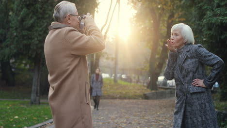 Anciano-Tomando-Una-Foto-Con-La-Cámara-De-Un-Smartphone-De-Su-Encantadora-Esposa-De-Pelo-Gris-En-El-Parque-En-Otoño