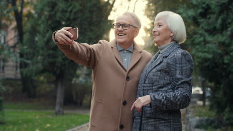 Elderly-Couple-Posing-And-Smiling-To-The-Camera-While-Taking-Selfie-Photo-With-Smartphone-In-The-Park-At-Sunset