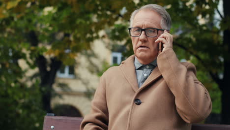 Elderly-Man-In-Glasses-And-Coat-Sitting-On-The-Bench-And-Talking-On-The-Phone-In-The-Park-In-Autumn