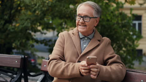 Älterer-Mann-Mit-Brille-Und-Mantel-Sitzt-Auf-Der-Bank-Und-Schreibt-SMS,-Während-Er-Im-Herbst-Im-Park-Auf-Das-Smartphone-Tippt