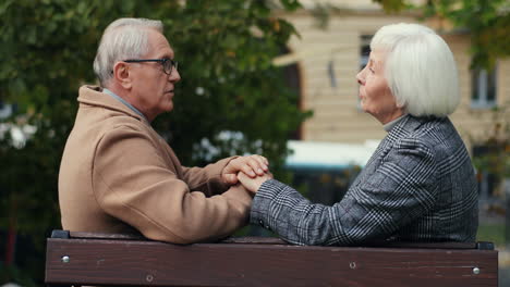 Side-View-Of-Elderly-Married-Couple-Holding-Hand-And-Talking-Sitting-On-The-Bench-In-The-Park-In-Autumn