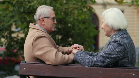 Side-View-Of-Elderly-Married-Couple-Holding-Hand-And-Talking-Sitting-On-The-Bench-In-The-Park-In-Autumn-1