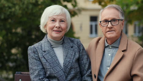 Porträt-Eines-älteren-Paares,-Das-Auf-Der-Bank-Sitzt-Und-Im-Herbst-Im-Park-In-Die-Kamera-Lächelt