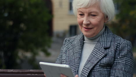 Close-Up-View-Of-Ederly-Woman-In-The-Coat-Sitting-On-The-Bench-In-The-Park-And-Tapping-Or-Typing-On-The-Tablet-In-Autumn