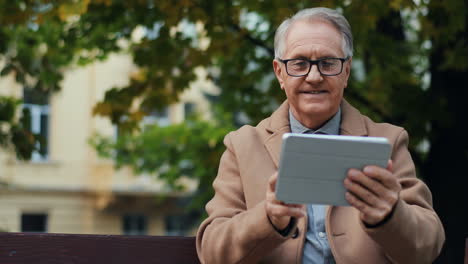 Elderly-Man-In-Glasses-And-Coat-Sitting-On-The-Bench-In-The-Park-And-Using-A-Tablet-In-Autumn