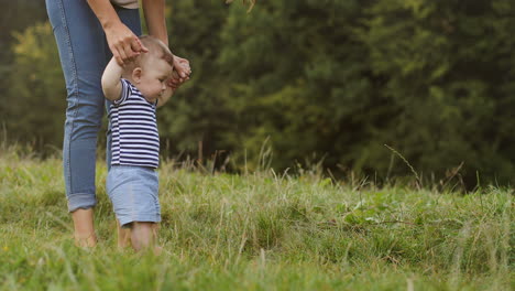 Cute-Little-Toddler-Boy-Holding-His-Mother's-Hands-And-Doing-First-Steps-Barefeet-On-The-Grass
