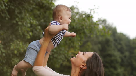 Portrait-Of-A-Beautiful-Young-Mother-Holding-And-Lifting-Her-Happy-Son-Up-In-The-Park