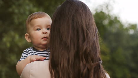 Back-View-Of-A-Woman-With-Long-Dark-Hair-Holding-Her-Smiling-Little-Son-Outdoors