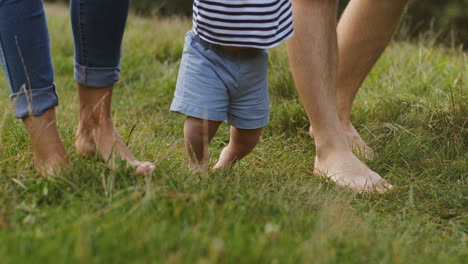 Close-Up-Of-A-Little-Baby-Taking-His-First-Steps-While-Walking-Barefoot-On-The-Green-Grass-And-Holding-His-Parents'-Hands