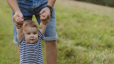 Close-Up-Of-A-Little-Baby-Taking-His-First-Steps-While-Walking-Barefoot-On-The-Green-Grass-And-Holding-His-Father's-Hands