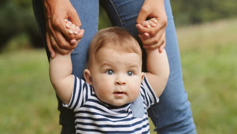 Close-Up-Of-A-Baby-Boy-Taking-His-First-Steps-In-The-Grass-While-Holding-His-Mother's-Hands