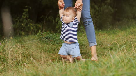 Portrait-Of-A-Baby-Boy-Taking-His-First-Steps-In-The-Grass-While-Holding-His-Mother's-Hands