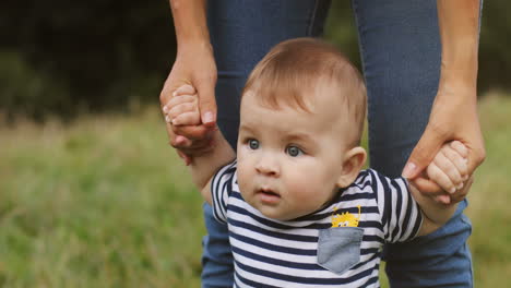Portrait-Of-A-Cute-Toddler-Boy-Standing-And-Holding-His-Mother's-Hands-Outdoors