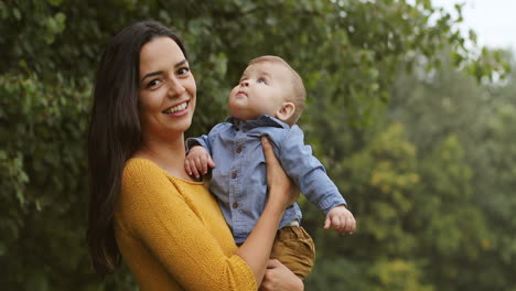 Portrait-Shot-Of-A-Smiling-Young-Woman-With-Long-Dark-Hair-Holding-Her-Baby-Boy-And-Looking-At-The-Camera-In-The-Park