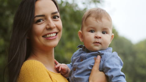 Portrait-Shot-Of-A-Smiling-Young-Woman-With-Long-Dark-Hair-Holding-Her-Cute-Baby-Boy-And-Looking-At-The-Camera-In-The-Park