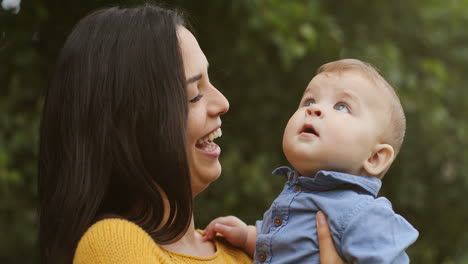 Portrait-Of-A-Cheerful-Mother-Holding-Her-Cute-Baby-Son-And-Smiling-At-The-Camera-In-The-Park