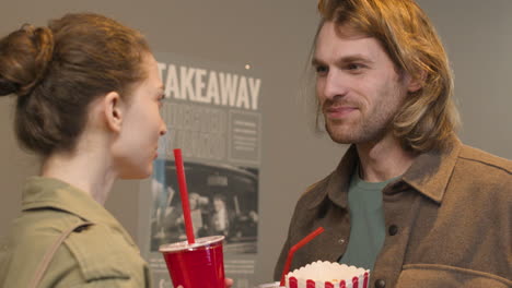 Happy-Couple-With-Popcorn-Box-And-Soft-Drinks-Talking-Together-And-Looking-A-Wall-Poster-At-The-Cinema