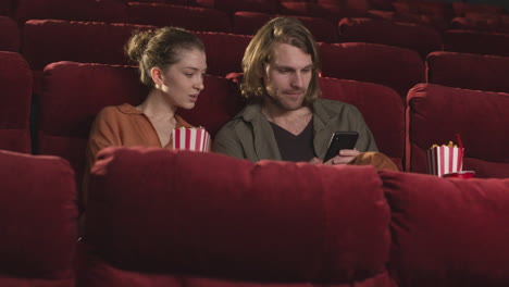 Happy-Couple-Sitting-In-Movie-Theater-And-Looking-Something-Funny-On-The-Mobile-Phone