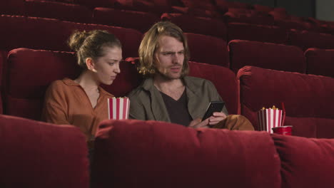 Blonder-Mann-Sitzt-Im-Kino-Und-Benutzt-Handy,-Während-Seine-Freundin-Popcorn-Isst