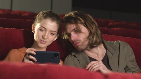 Glückliches-Paar,-Das-Auf-Dem-Handy-Etwas-Interessantes-Sieht,-Während-Es-Vor-Dem-Filmstart-Im-Kino-Sitzt
