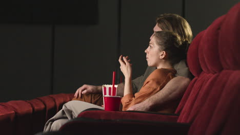 Side-View-Of-Couple-Hugging-Sitting-In-The-Cinema-While-They-Watching-A-Movie-And-Eating-Popcorn