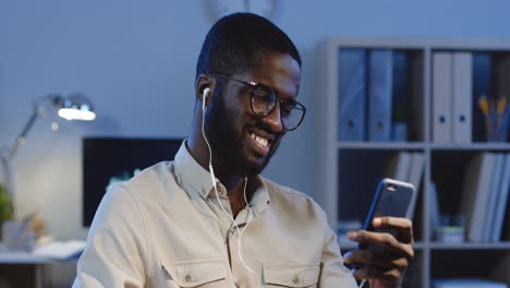 Close-Up-View-Of-Young-Man-In-Glasses-And-Headphones-Listening-To-The-Music-On-Smartphone-And-Nodding-Head-In-Rhythm-In-The-Office-At-Night