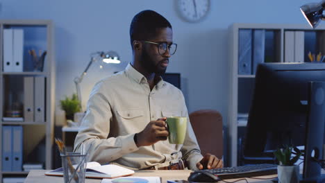 Young-Man-Drinking-Coffee-While-Working-At-The-Computer-In-The-Office-At-Night