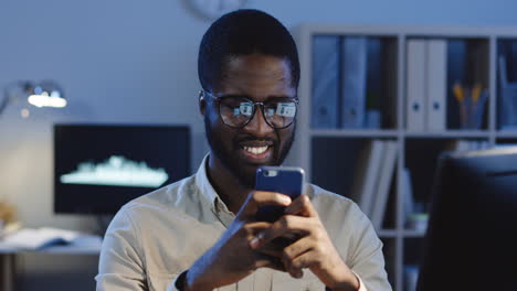 Portrait-Of-Young-Smiled-Office-Worker-Wearing-Glasses-Typing-And-Chatting-On-The-Smartphone-In-The-Office-At-Night