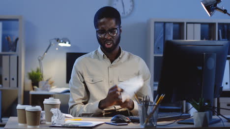 Angry-Young-Man-In-Glasses-Working-With-Computer-And-Writing-Something,-Then-Throws-A-Sheet-Of-Paper-Away-In-The-Office-At-Night