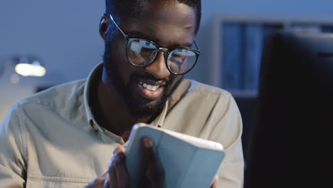 Close-Up-View-Of-Young-Cheerful-Man-In-Glasses-Using-His-Tablet-In-The-Office-At-Night